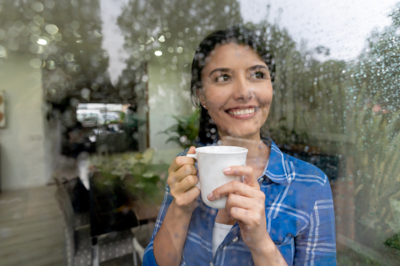 Beautiful woman at home looking through the window on a rainy day enjoyng a cup of coffee 
