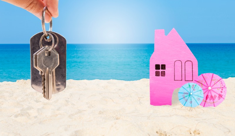 Miniature pink wooden house and hand holding key on tropical beach